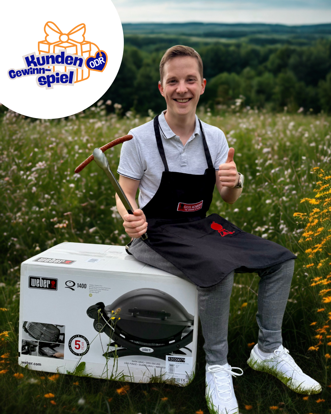 Ein junger Mann mit Grillschürze und Rostbratwurst in einer Zange sitzt auf der Verpackung eines Weber Elektrogrills vor eine Blühwiese.