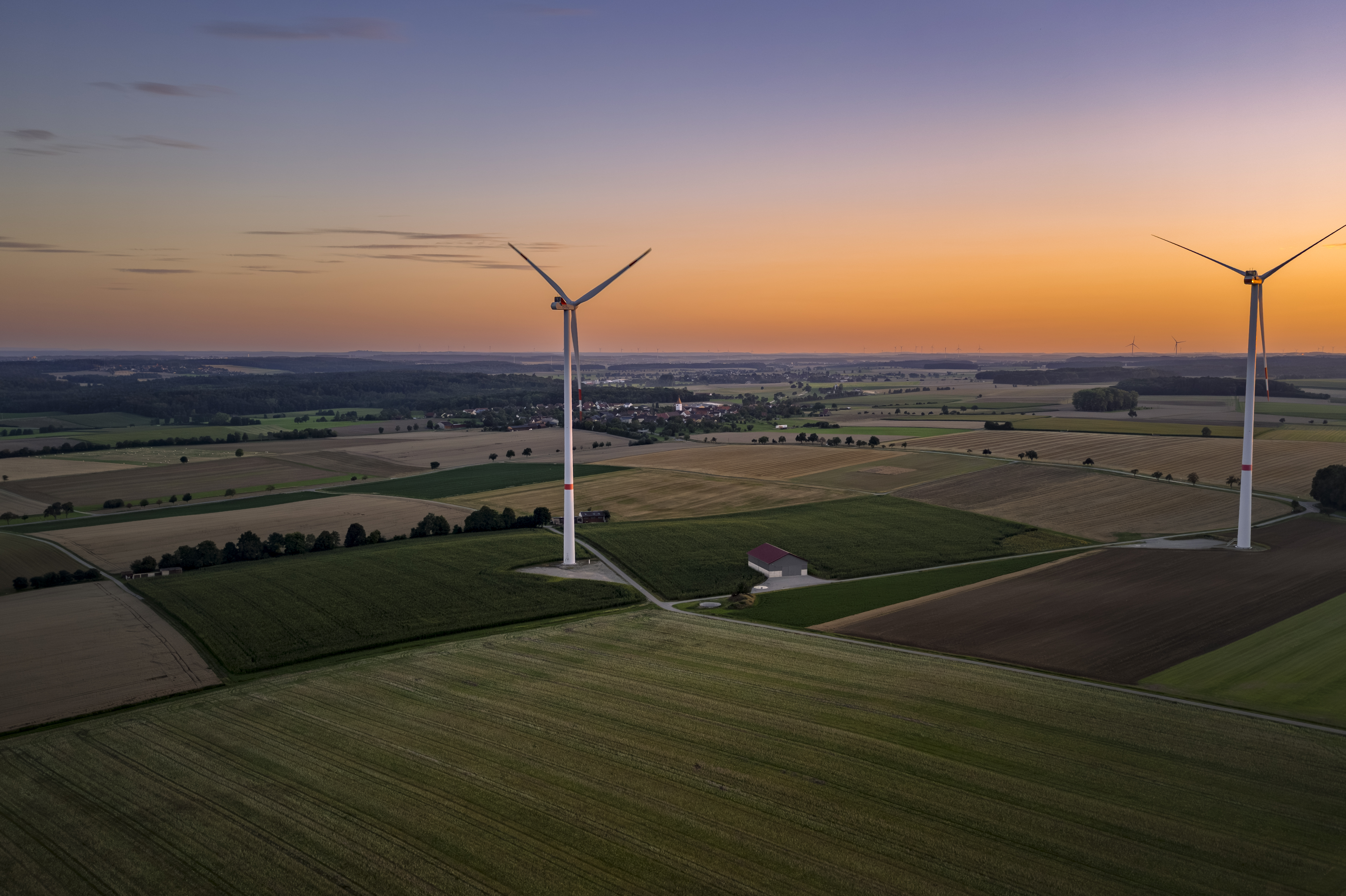 Zwei Windräder stehen auf Feldern bei abendlicher Stimmung mit Sonnenuntergang.