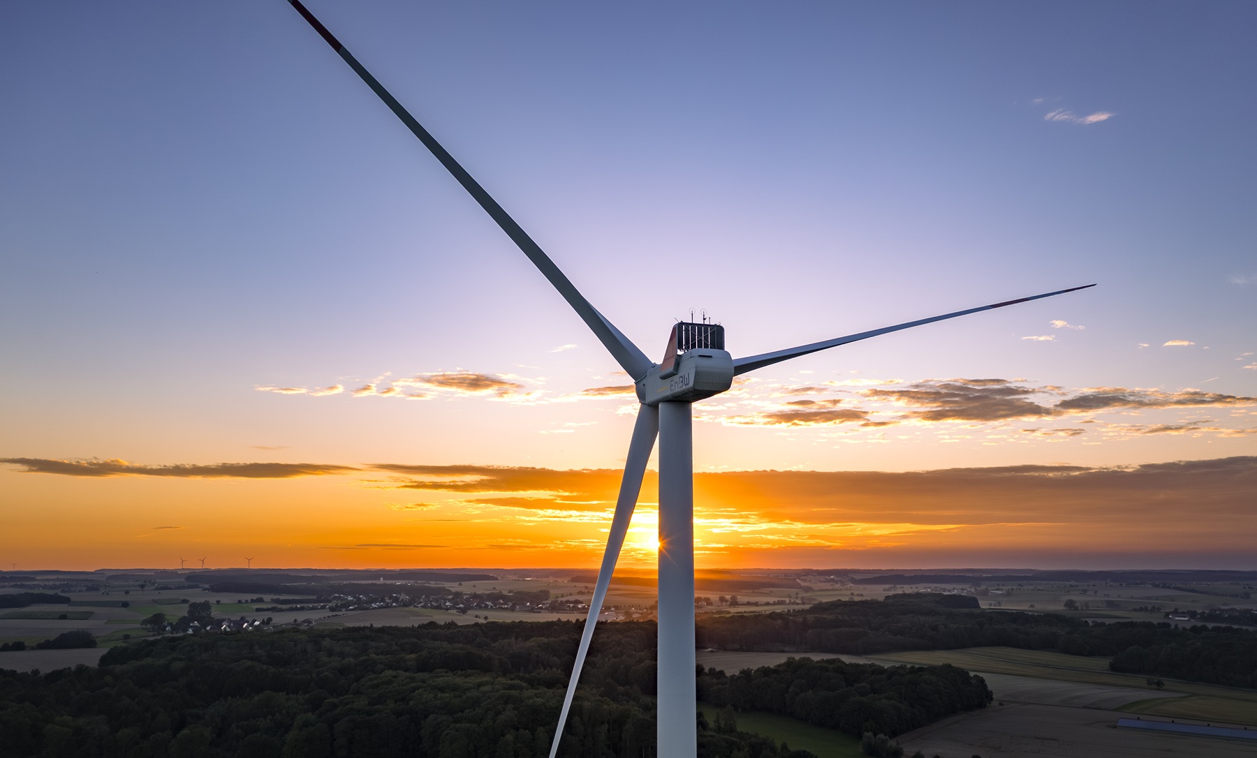 Ein fokussiertes Windrad mit drei Rotorblättern bei Sonnenuntergang. Im Hintergrund ist eine Landschaft zu sehen.