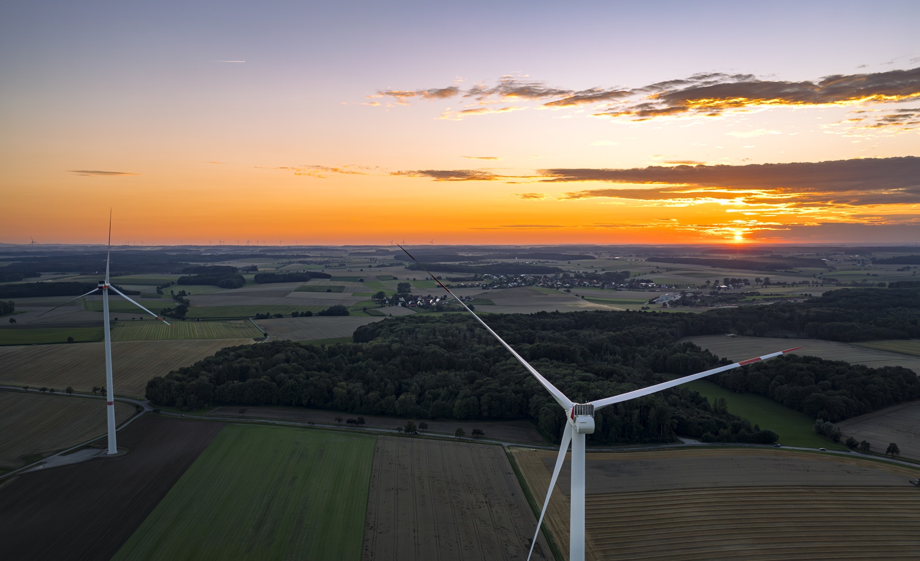 Zwei Windräder von der Rückseite bei Sonnenuntergang in einer grünen Landschaft mit Wiesen und Wäldern.