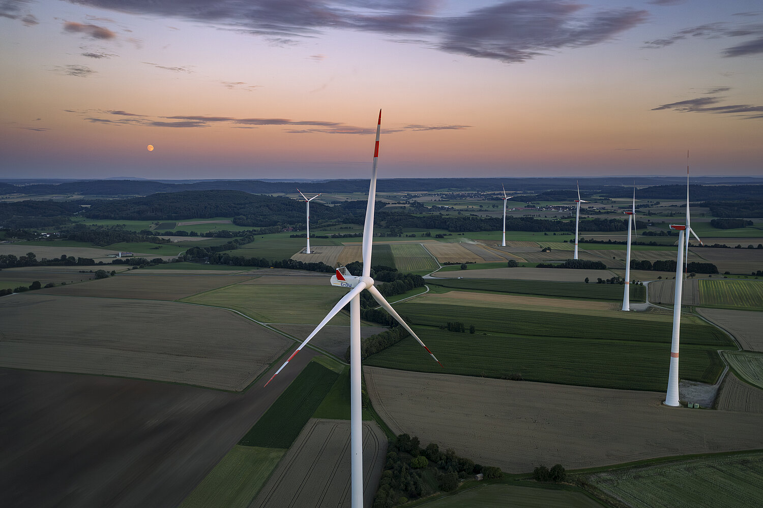Ein Windpark mit fünf Windrädern bei Sonnenuntergang in einer grünen Wiesenlandschaft.