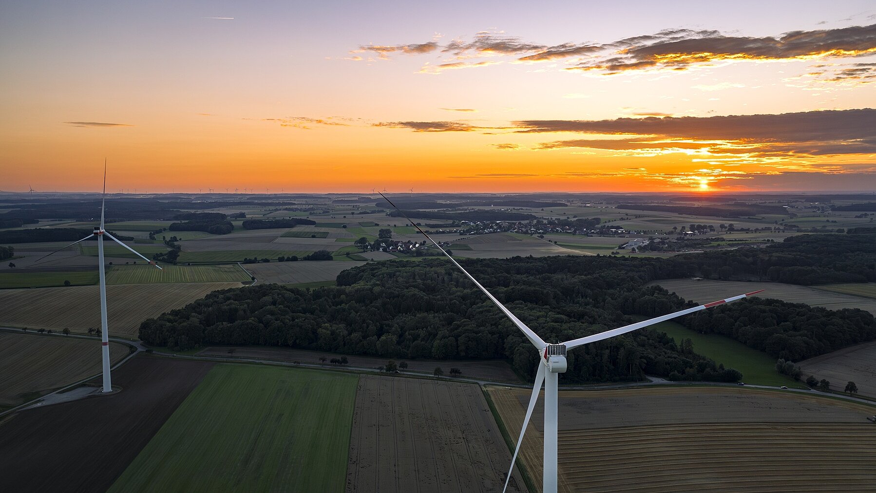 Zwei Windräder von der Rückseite bei Sonnenuntergang in einer grünen Landschaft mit Wiesen und Wäldern.