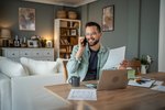 Ein telefonierender Kunde sitzt in seiner Wohnung am Tisch mit einem Laptop und Papierunterlagen.