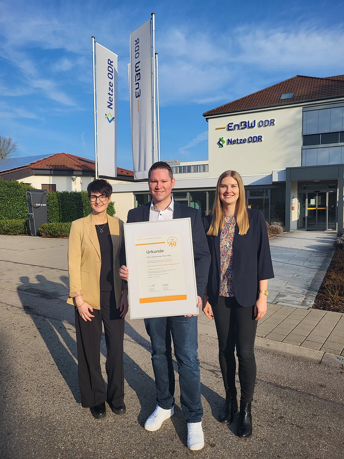 Drei Personen stehen vor einem Gebäude mit den Logos „EnBW ODR“ und „Netze ODR“. Die mittlere Person hält eine große Urkunde im Hochformat. Hinter der Gruppe wehen zwei Flaggen mit dem Schriftzug "EnBW ODR" und „Netze ODR“. Die Szene ist im Außenbereich bei sonnigem Wetter aufgenommen.