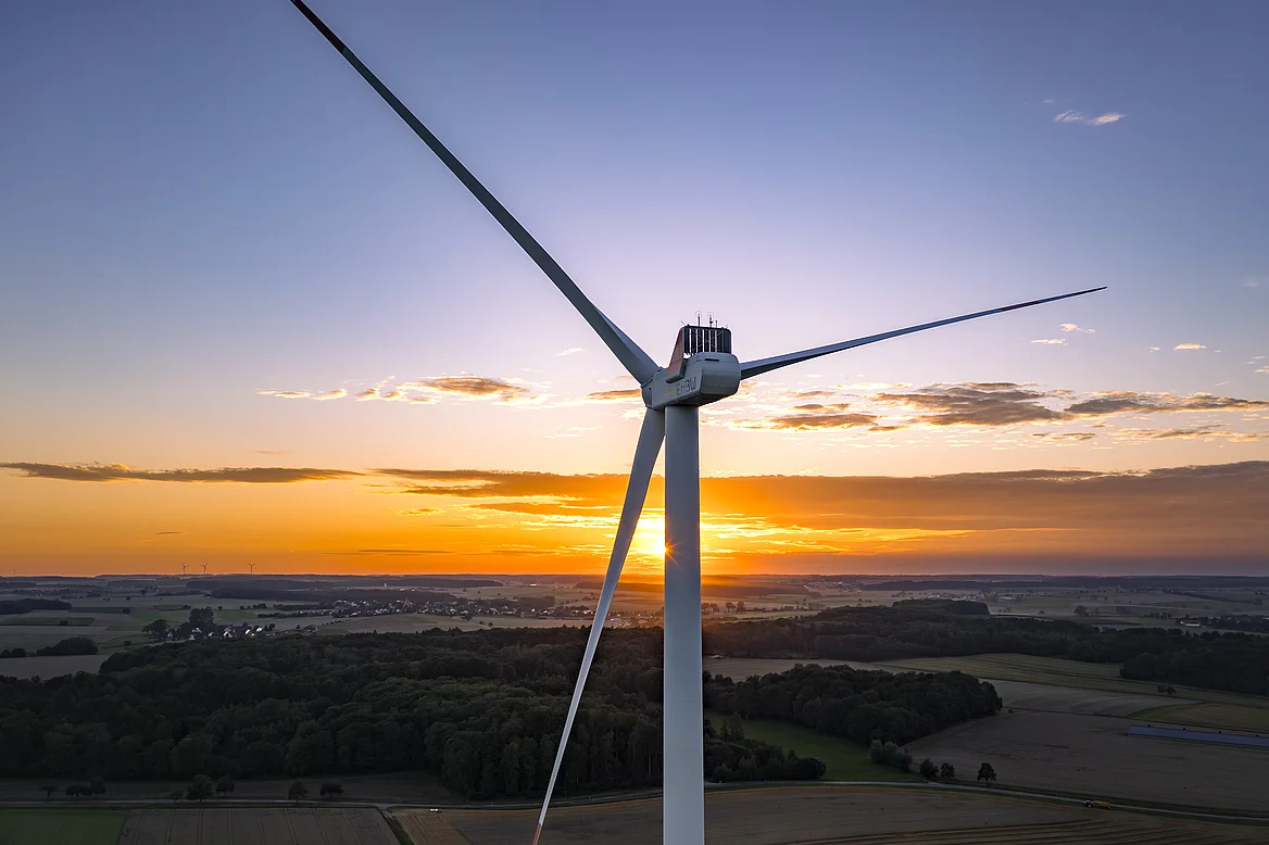 Ein fokussiertes Windrad mit drei Rotorblättern bei Sonnenuntergang. Im Hintergrund ist eine Landschaft zu sehen.