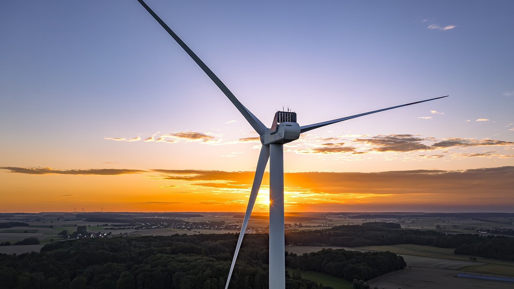 Ein fokussiertes Windrad mit drei Rotorblättern bei Sonnenuntergang. Im Hintergrund ist eine Landschaft zu sehen.