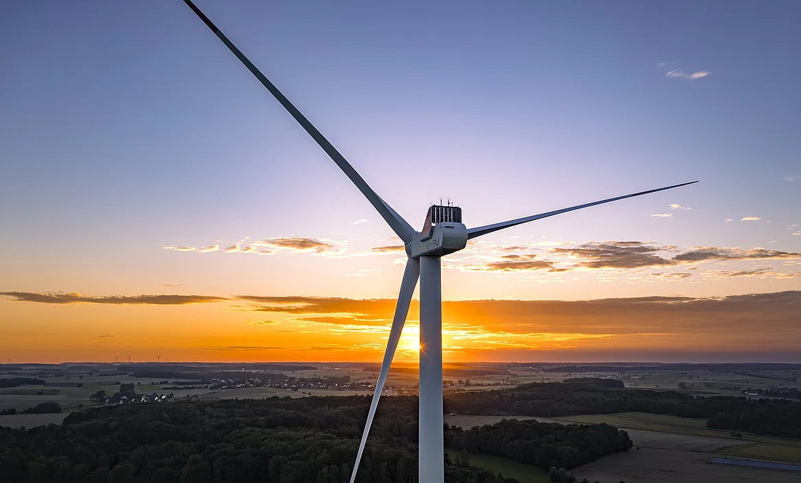 Ein fokussiertes Windrad mit drei Rotorblättern bei Sonnenuntergang. Im Hintergrund ist eine Landschaft zu sehen.