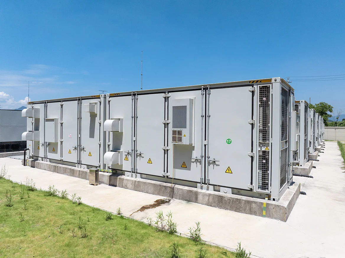 Großer Batteriespeicher aus weißen Container-Modulen, die bei strahlendem Sonnenschein auf einer grünen Fläche stehen, vor blauem Himmel und in natürlicher Umgebung.