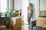 Eine Kundin lehnt zu Hause an der Wand ihrer neuen Wohnung umgeben von Umzugskartons und informiert sich über ihr Smartphone.