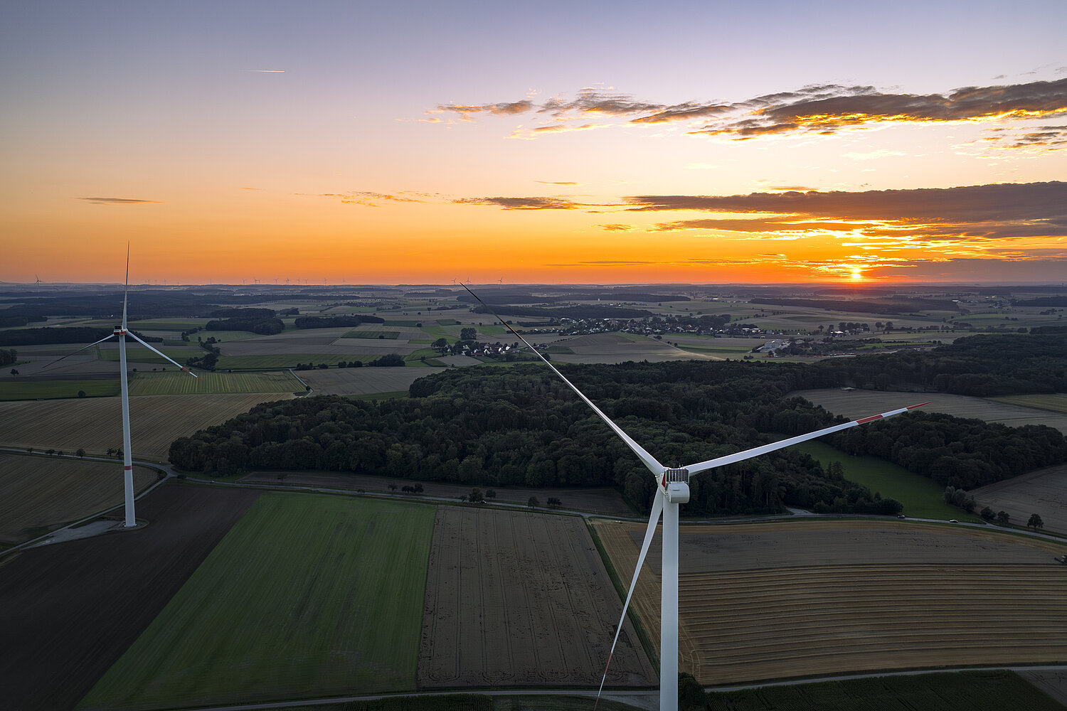 Zwei Windräder von der Rückseite bei Sonnenuntergang in einer grünen Landschaft mit Wiesen und Wäldern.