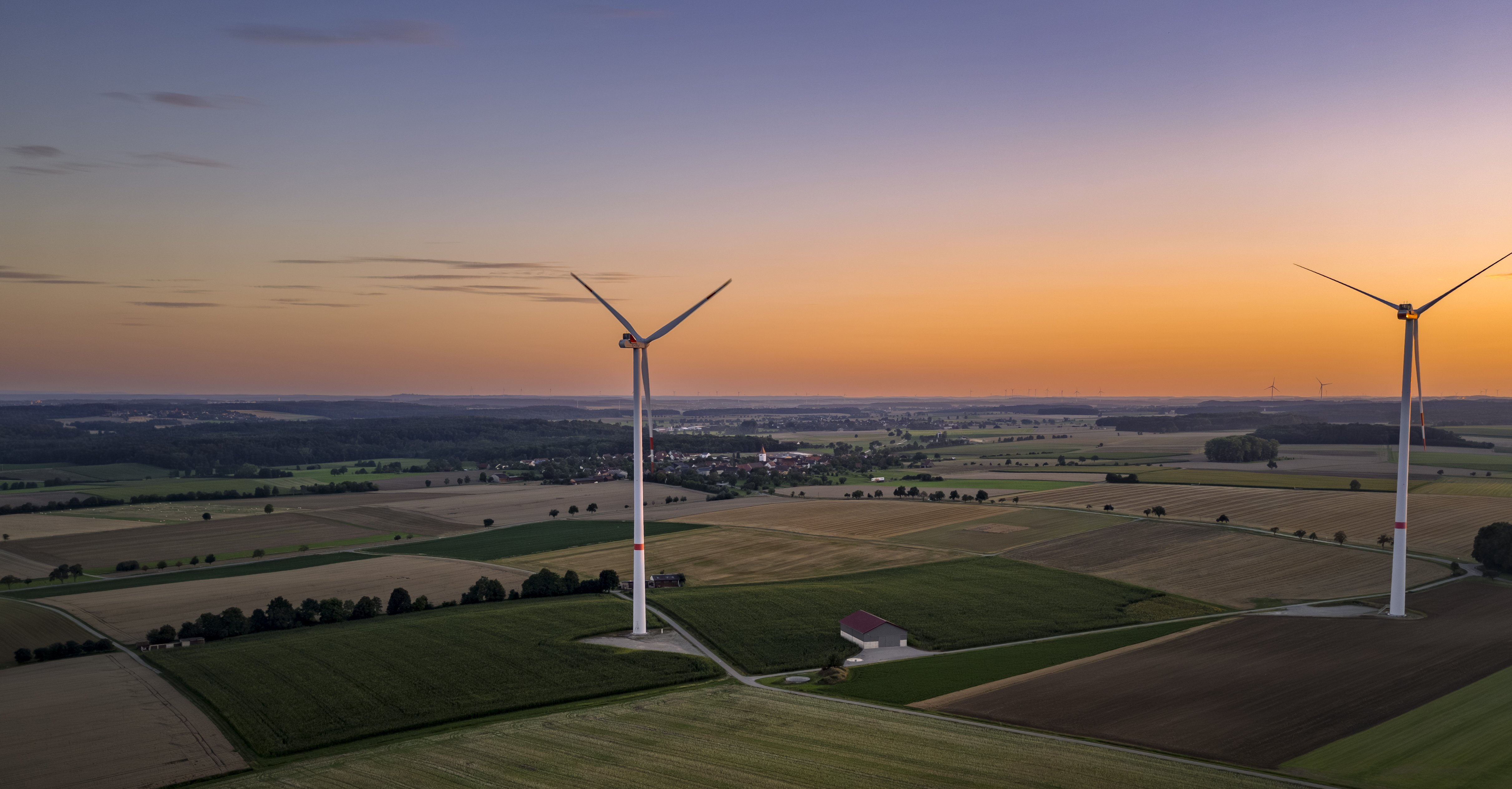 Zwei Windräder stehen auf Feldern bei abendlicher Stimmung mit Sonnenuntergang.