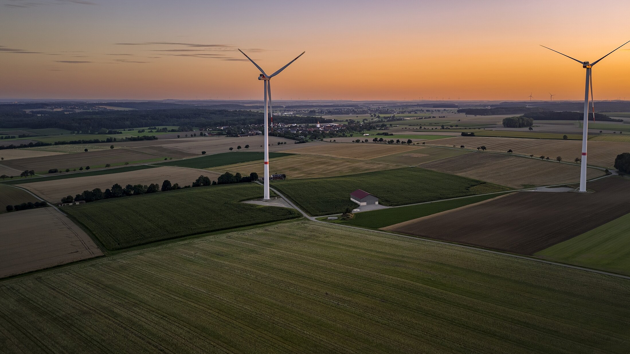 Zwei Windräder stehen auf Feldern bei abendlicher Stimmung mit Sonnenuntergang.